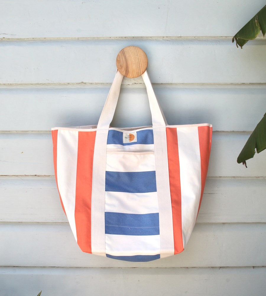 Striped tote bag with red, white, and blue pattern hanging on a wooden hook against a light wooden panel background.