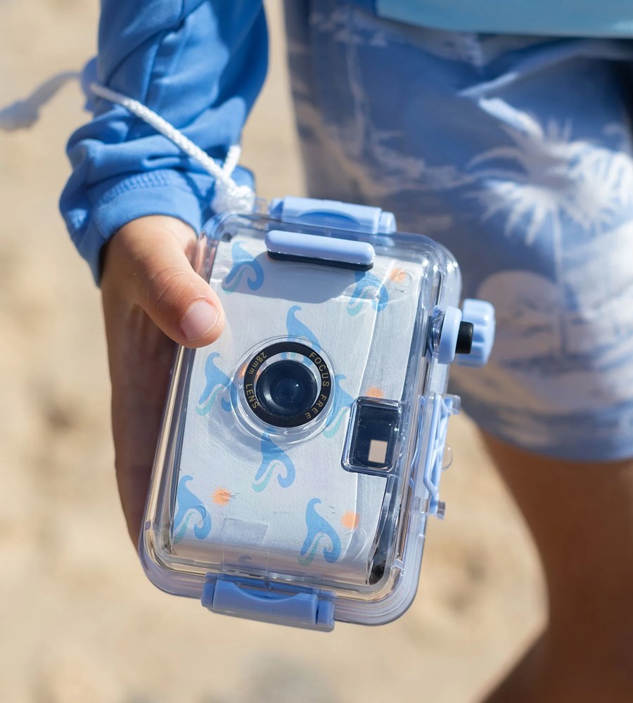 Clear camera with blue accents held by a person in a blue jacket and patterned shorts.