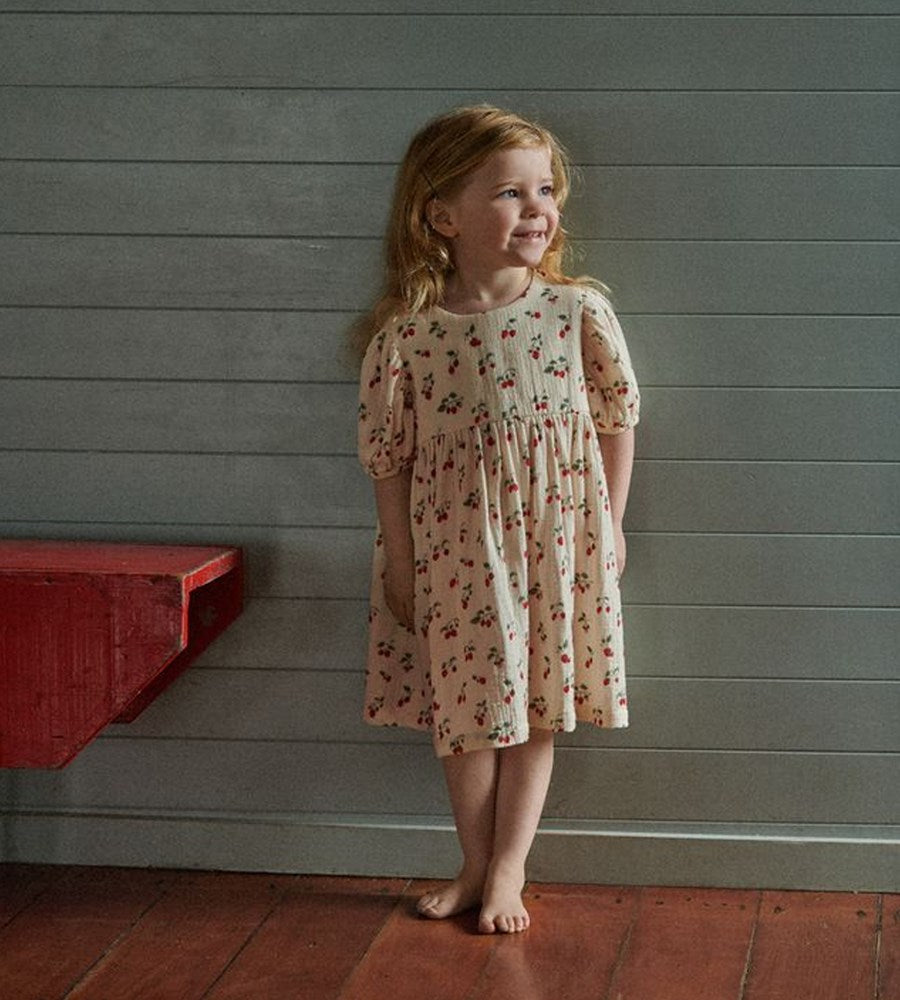 Young girl in a floral dress standing against a gray wall with a red shelf.