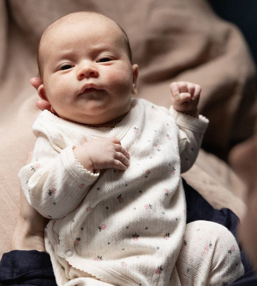 Baby in a white outfit with small patterns, sitting on a person's lap.