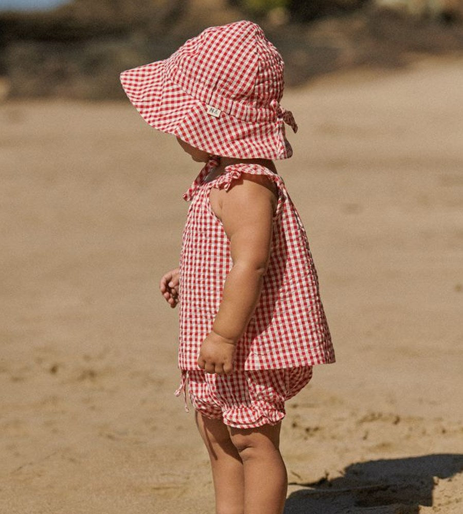 Child wearing a red and white checkered dress and sun hat on a sandy beach.