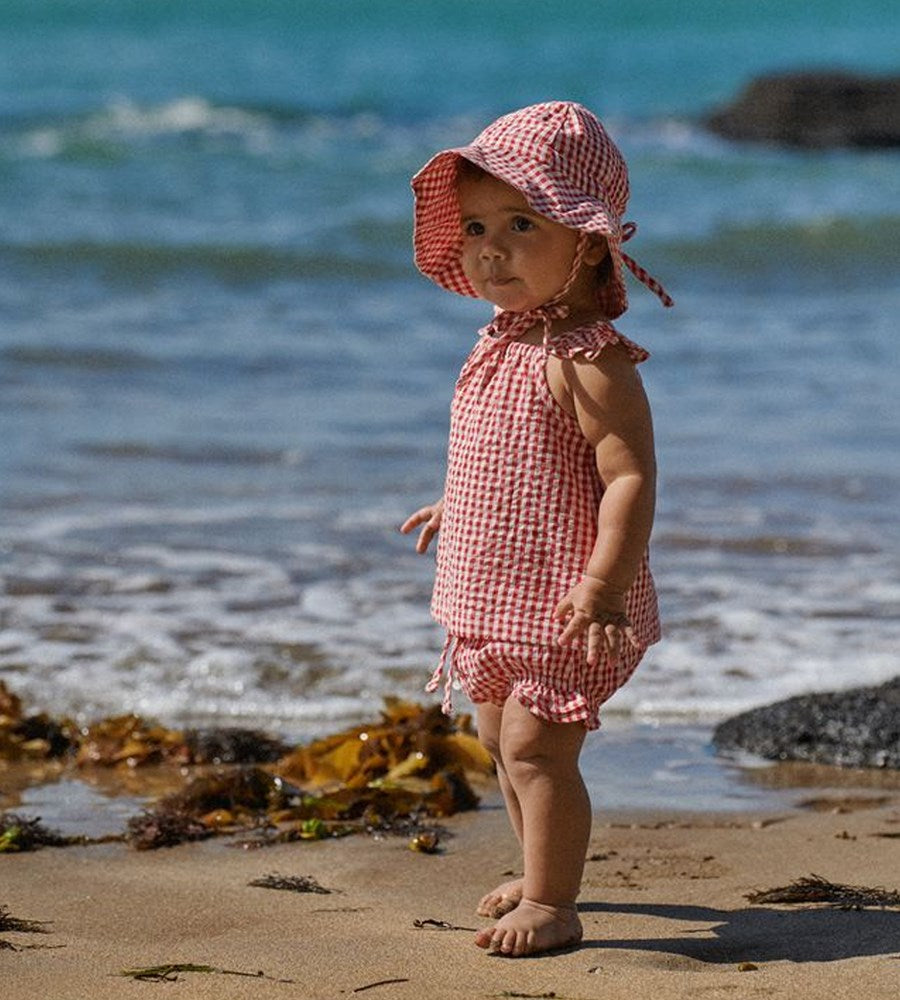 Child in a red checkered outfit and hat standing on a beach with ocean in the background