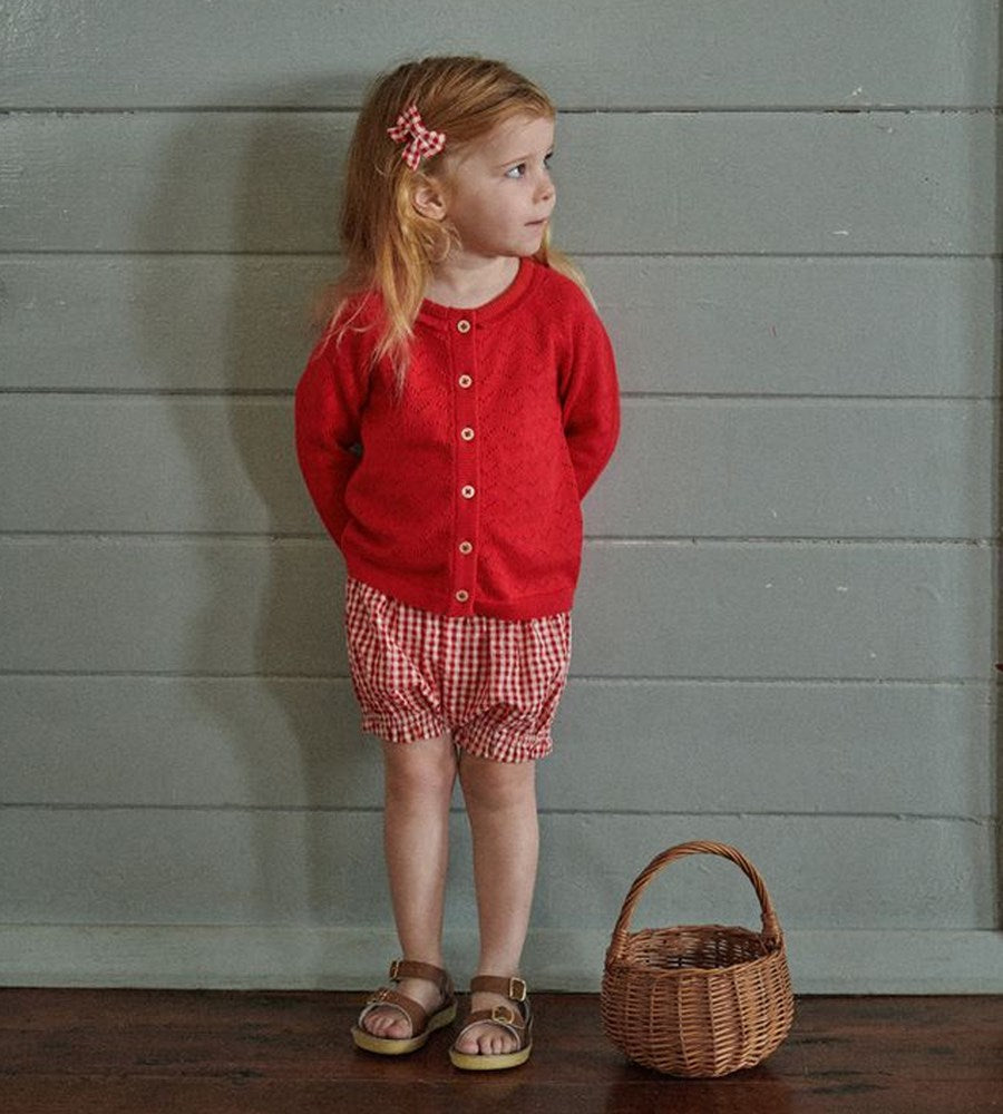 Young girl in a red cardigan and checkered shorts standing next to a wicker basket against a wooden paneled wall.