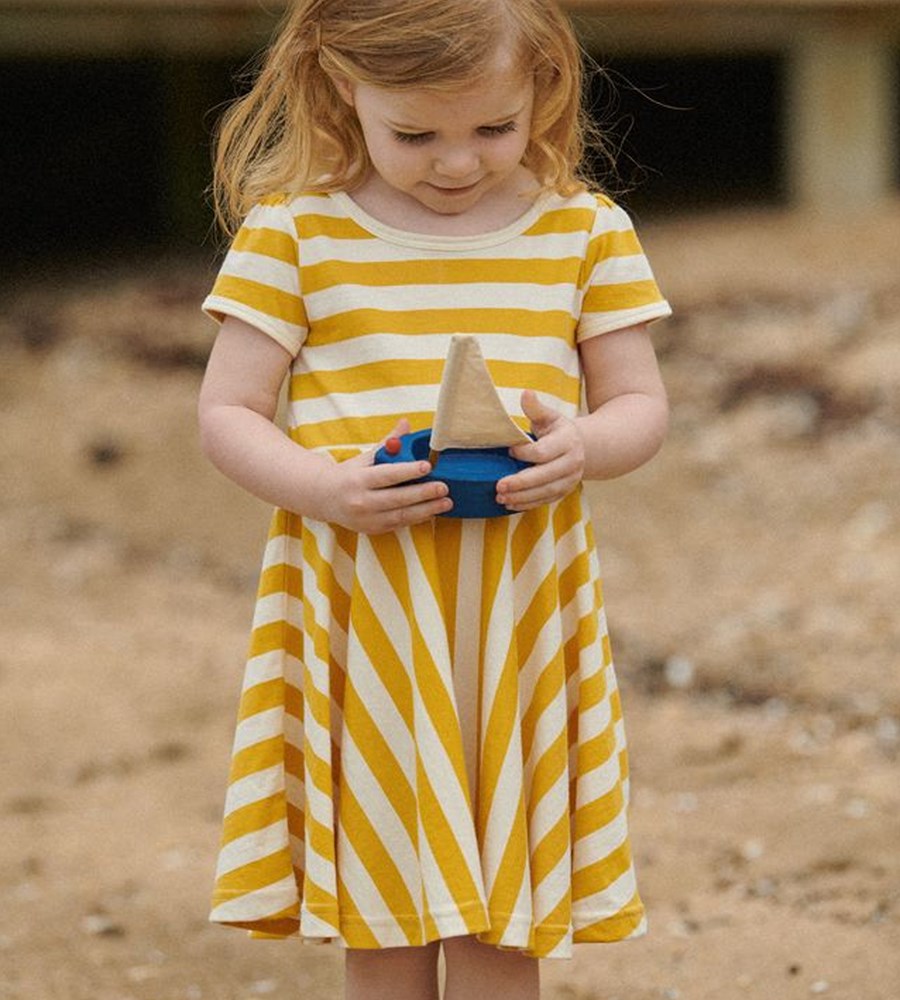 Young girl in a yellow and white striped dress holding a toy on a sandy ground.