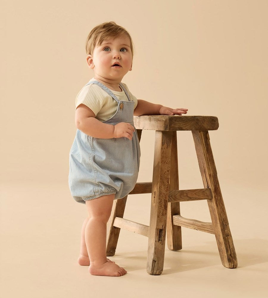 Child in denim overalls standing next to a wooden stool on a beige background