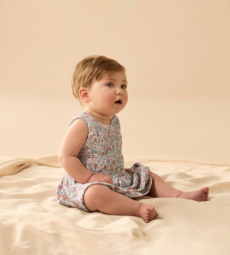 Baby sitting on a beige blanket wearing a floral dress.