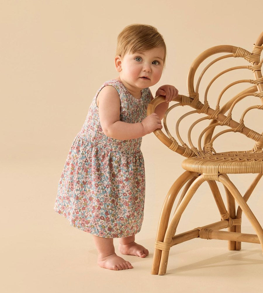 Baby in a floral dress standing next to a wicker chair on a beige background