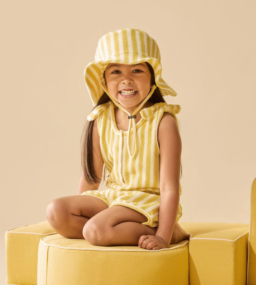 Child wearing a yellow striped outfit and sun hat sitting on a yellow chair against a beige background